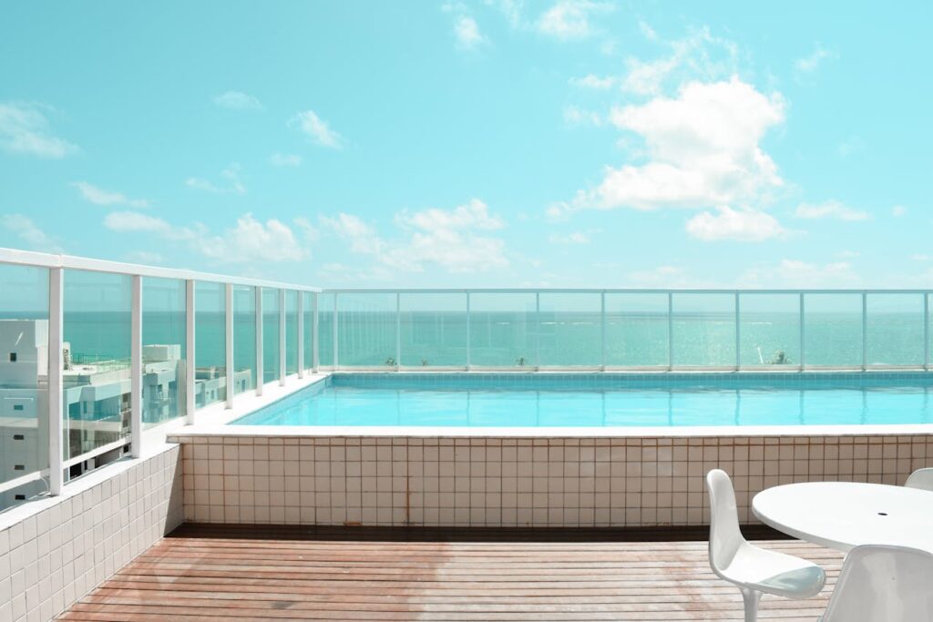 A serene rooftop infinity pool with glass railings overlooking the blue ocean under a partly cloudy sky.