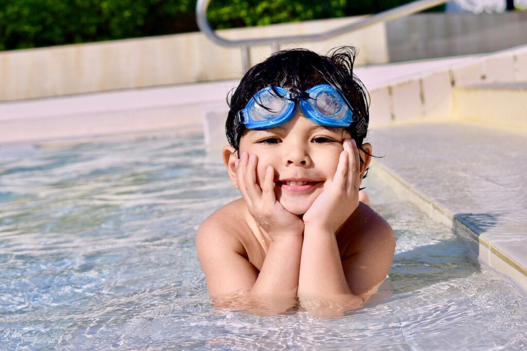 A smiling child with blue swimming goggles rests their head on their hands in a shallow pool.